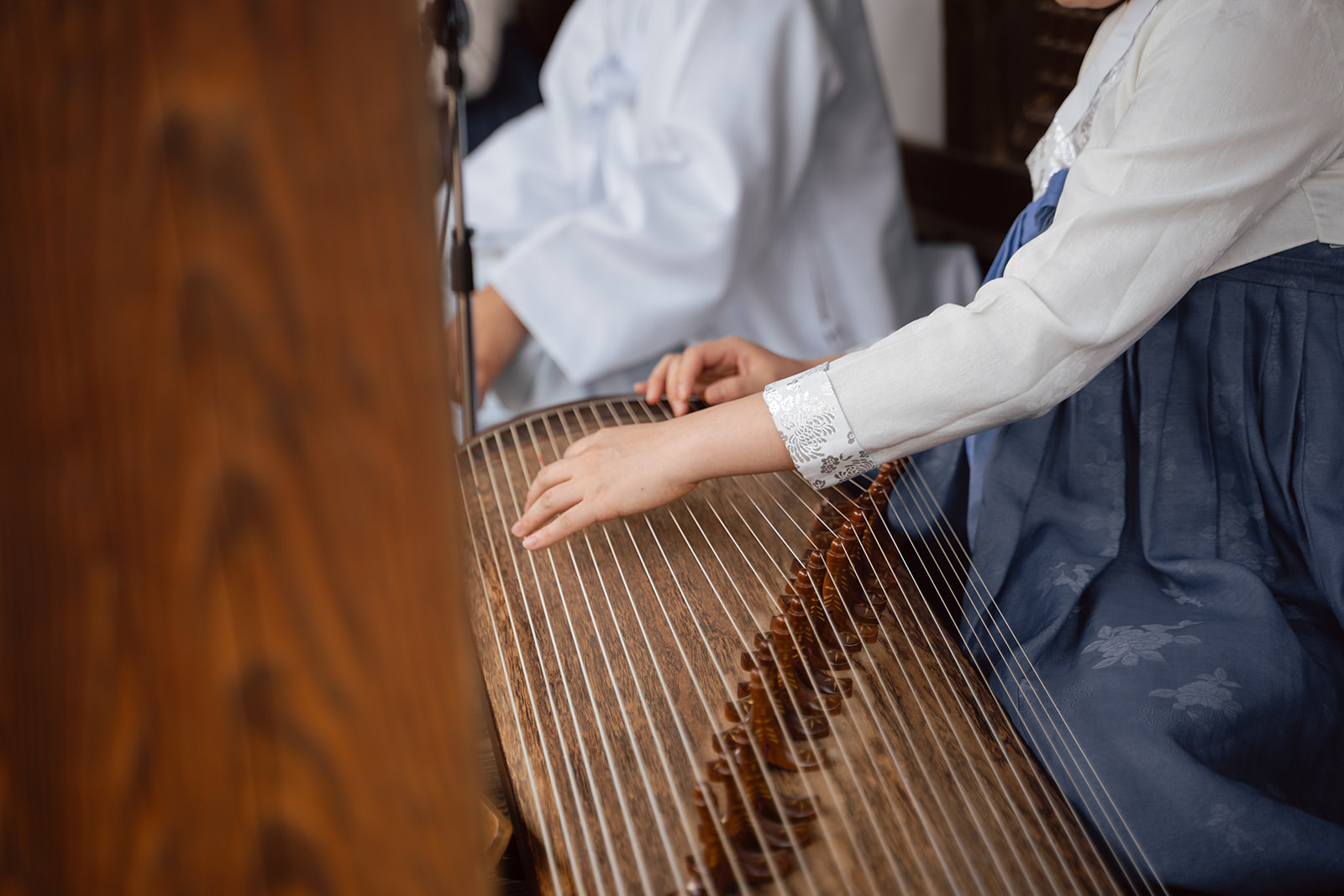 A person wearing traditional clothing plays a stringed instrument, possibly a gayageum, with both hands.
