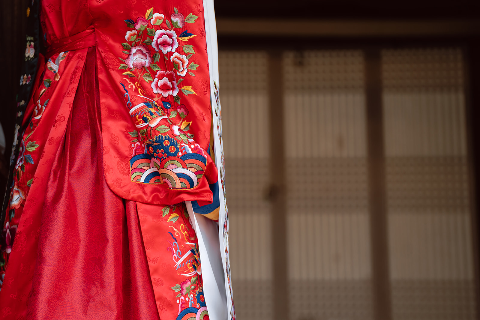 Close-up of a person wearing a traditional red garment with colorful floral embroidery, perfect for a Korea wedding day, standing in front of a blurred, patterned background.