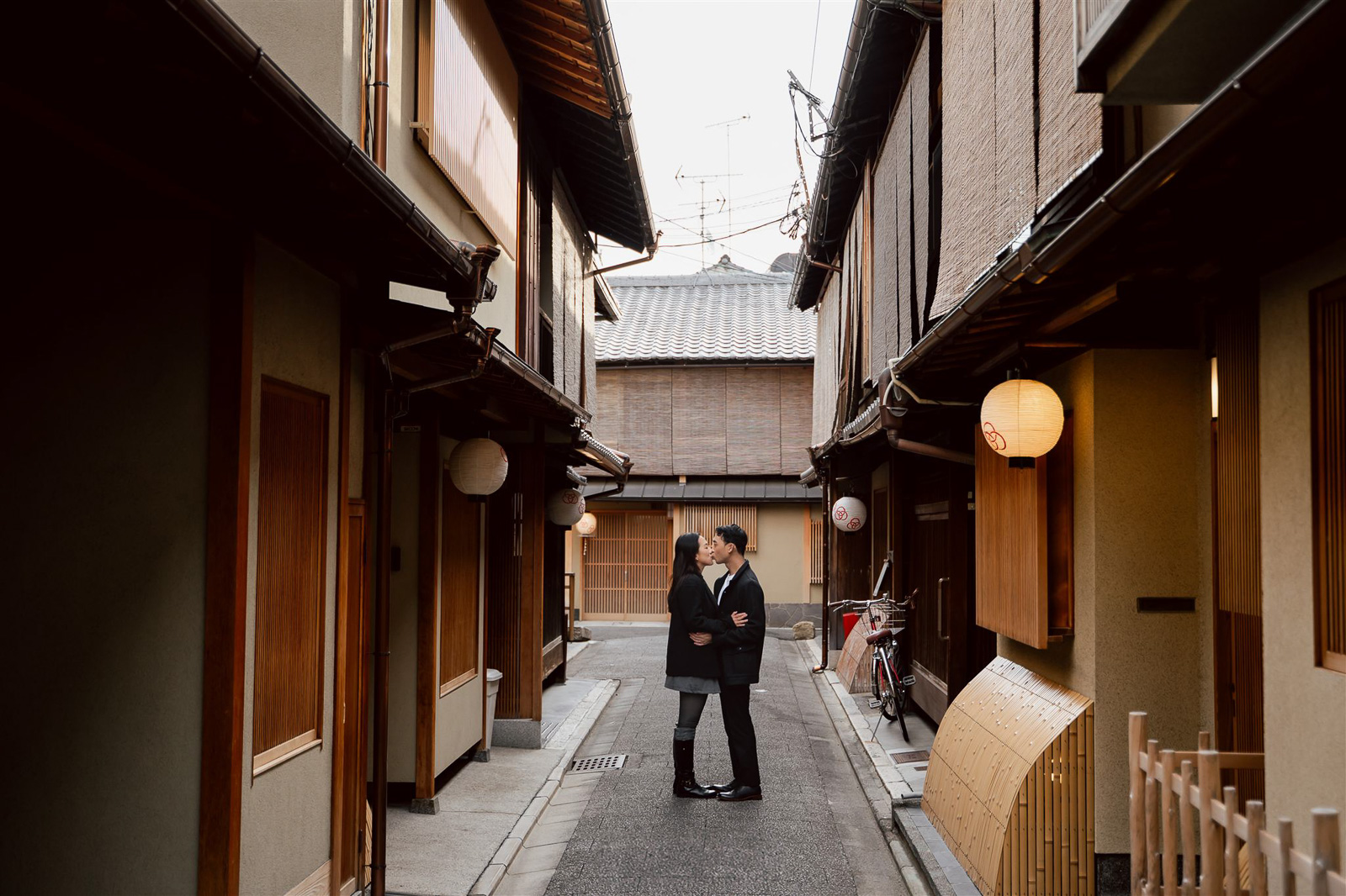 A couple stands closely together, facing each other in a narrow alleyway between traditional wooden buildings with paper lanterns—perfect for capturing intimate wedding photos in Japan.