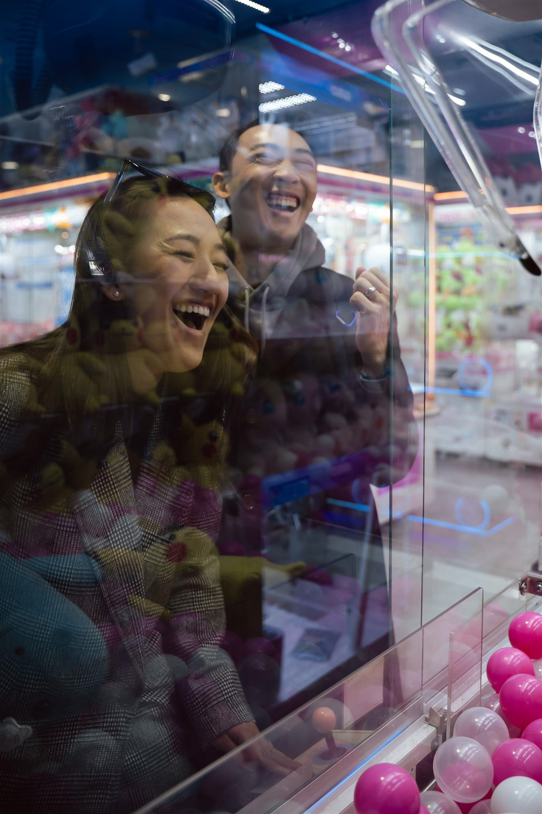 Two people smiling and laughing while playing a claw machine game in an arcade, surrounded by colorful balls.