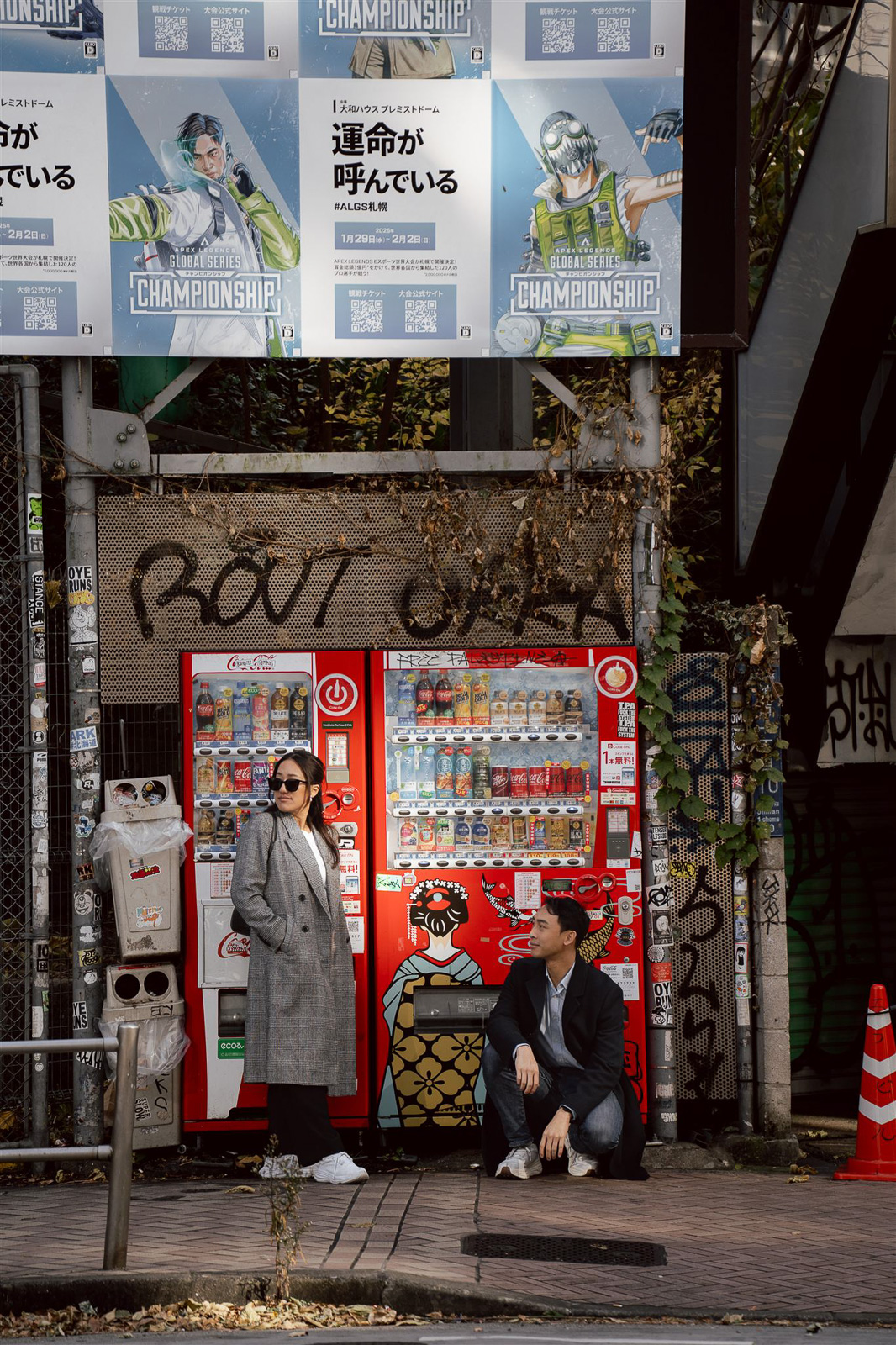 Two people pose in front of red vending machines on a city street, surrounded by posters, graffiti, and urban greenery—capturing the candid spirit often seen in unique wedding photos in Japan.