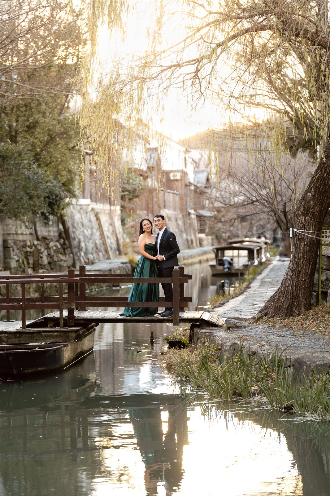 A couple stands on a narrow wooden bridge over a canal, with trees, boats, and old stone buildings in the background at sunset—capturing the magic of wedding photos in Japan.
