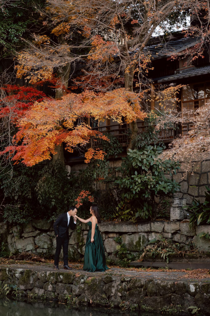 A man in a suit bows to a woman in a green dress by a stone path, with autumn trees and a traditional wooden building—perfect for capturing elegant wedding photos in Japan.