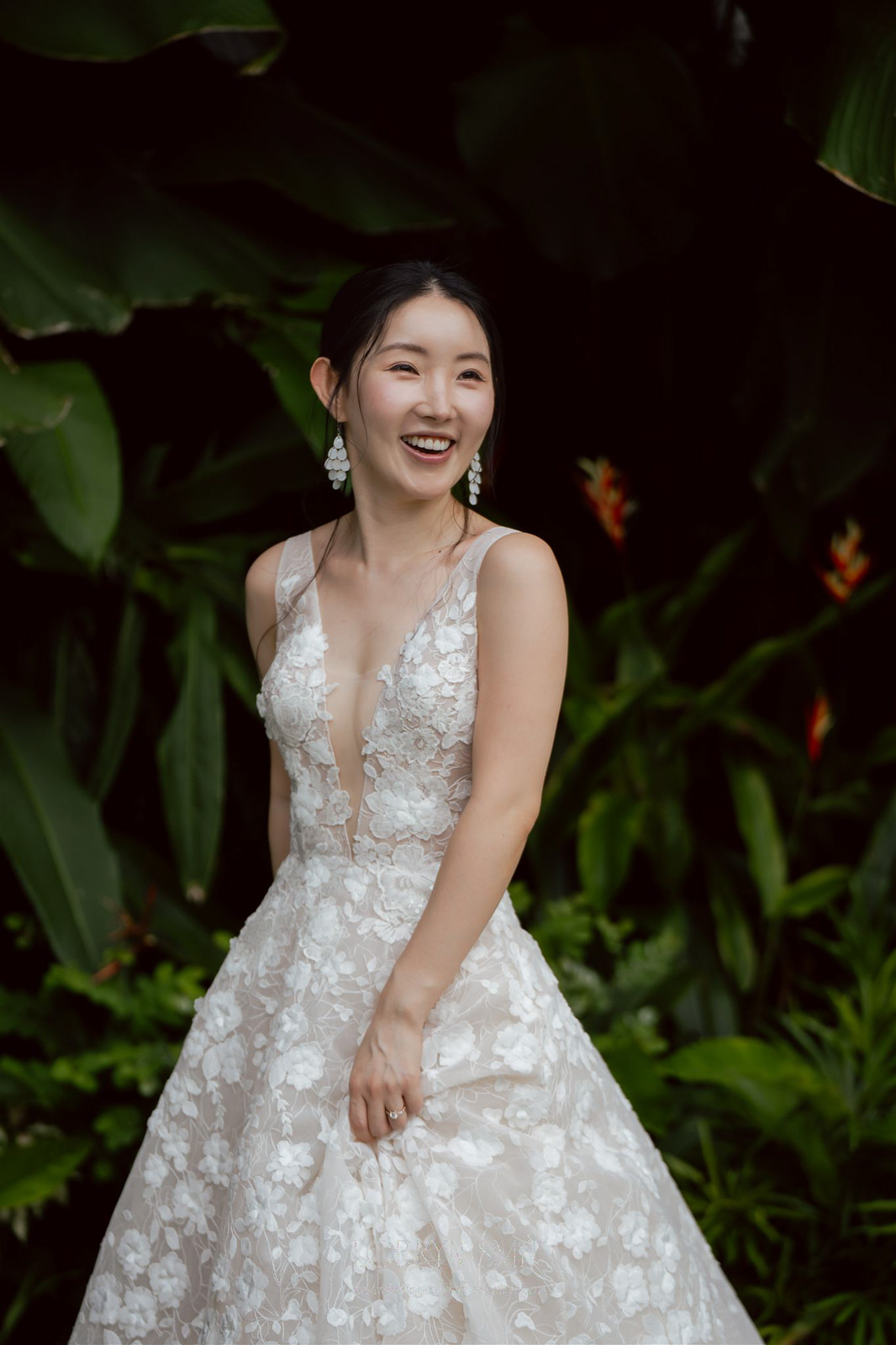 A woman in a white, floral-patterned wedding dress stands outdoors, smiling, with green foliage and flowers in the background.