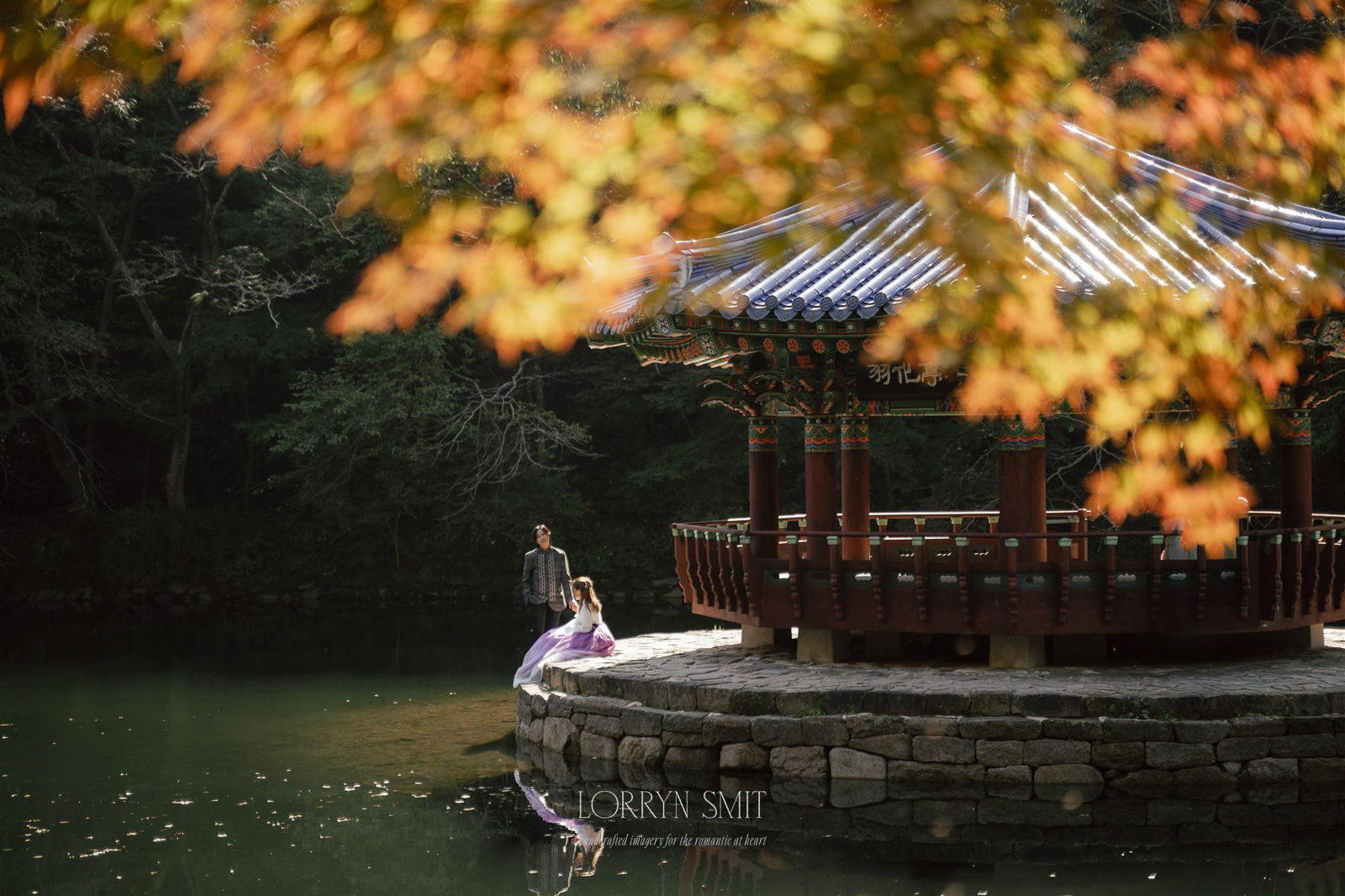 A man and woman stand near a traditional pavilion beside a pond, surrounded by trees with autumn foliage—an enchanting scene reminiscent of top Asia wedding destinations.