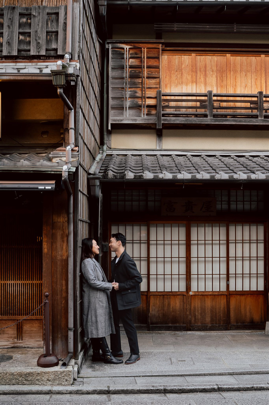 A couple stands close together, facing each other, on a quiet street in front of traditional wooden Japanese buildings—one of Asia's enchanting wedding destinations.