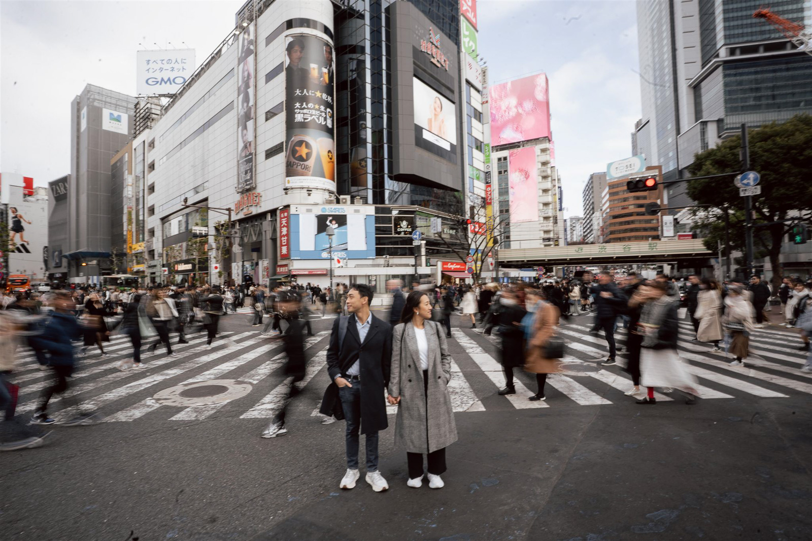 A couple stands holding hands in the middle of a busy crosswalk while people blur past them in an urban city setting with tall buildings and billboards, capturing the romance found in top Asia wedding destinations.