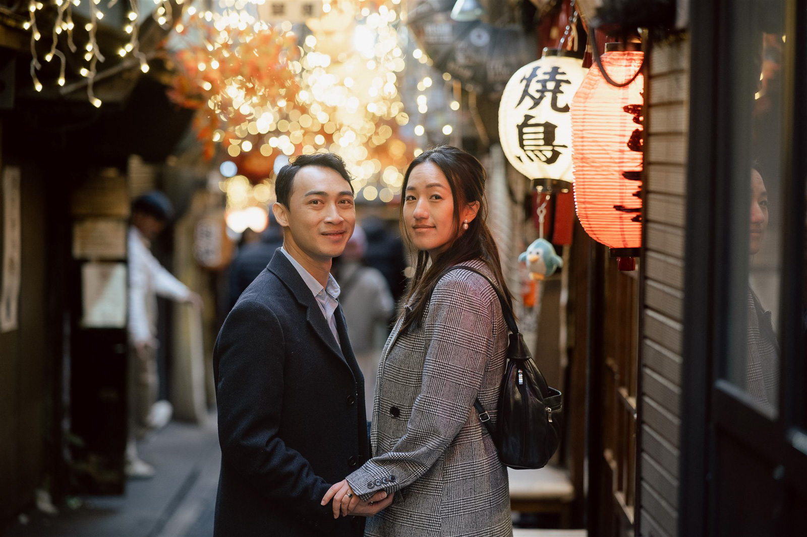 A couple stands closely together in a narrow, lantern-lit alleyway decorated with string lights and signs, evoking the charm of Asia wedding destinations as they look toward the camera.