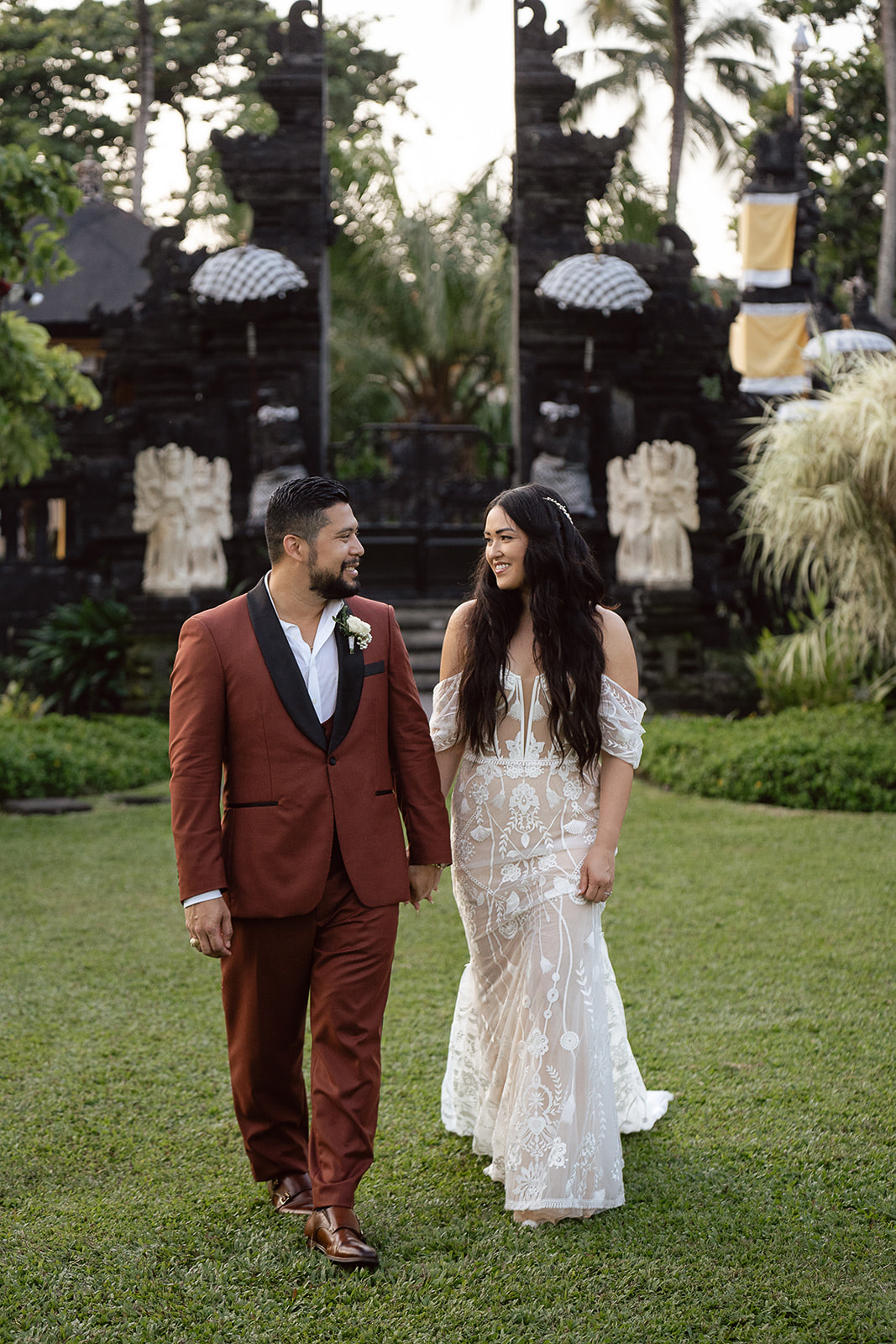 A couple dressed in wedding attire walks on grass before traditional black stone gates and lush greenery, capturing the enchanting beauty of Asia wedding destinations.