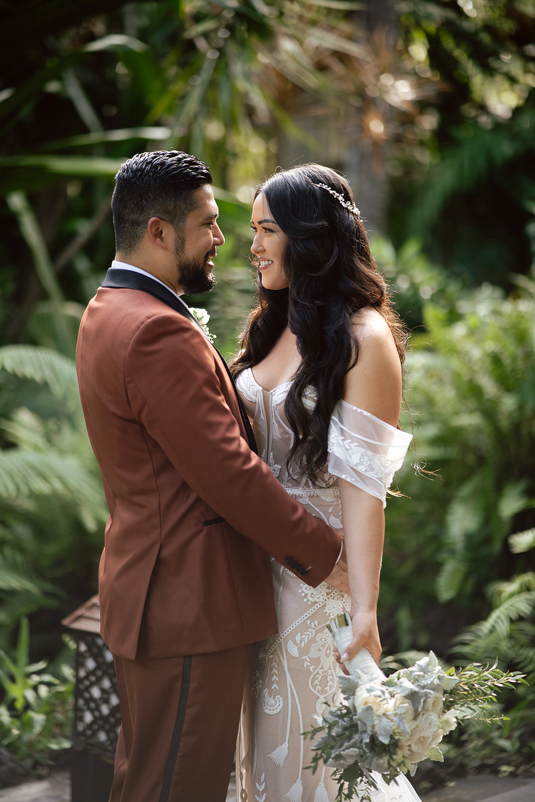 A couple in formal wedding attire stands close together outdoors, smiling at each other. The bride holds a bouquet and the groom wears a brown suit, surrounded by lush greenery reminiscent of popular Asia wedding destinations.