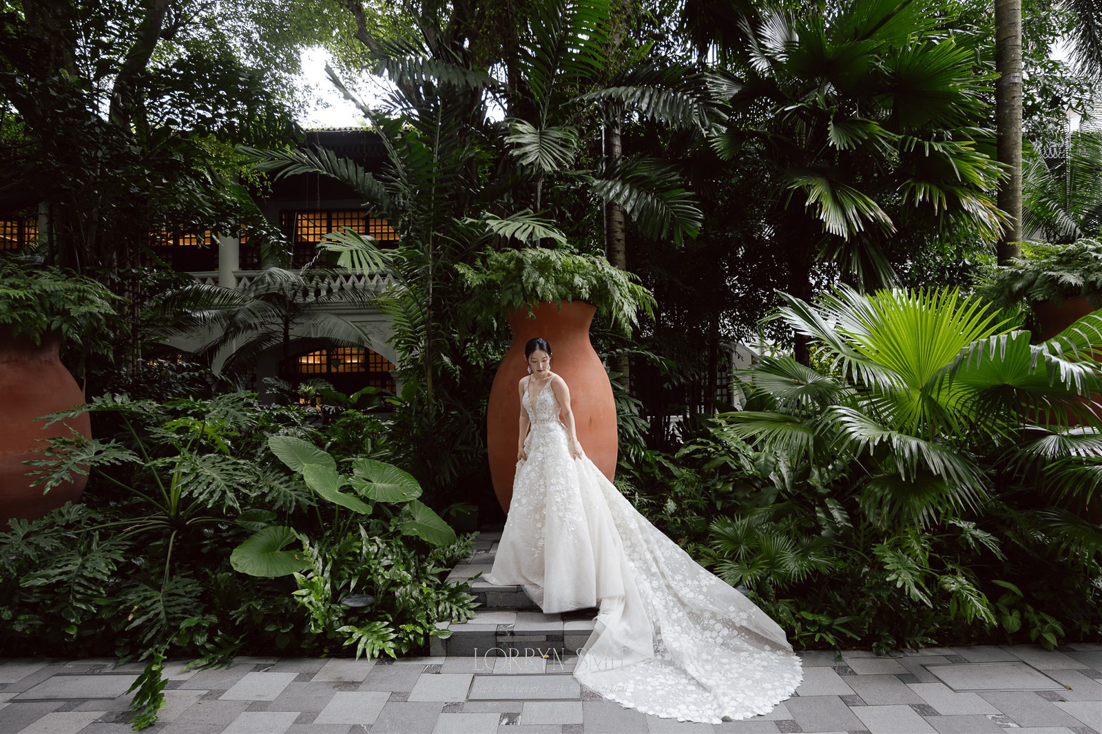 A bride in a long white wedding gown stands on a stone path surrounded by lush green tropical plants and large terracotta pots, evoking the enchanting beauty of Asia wedding destinations.