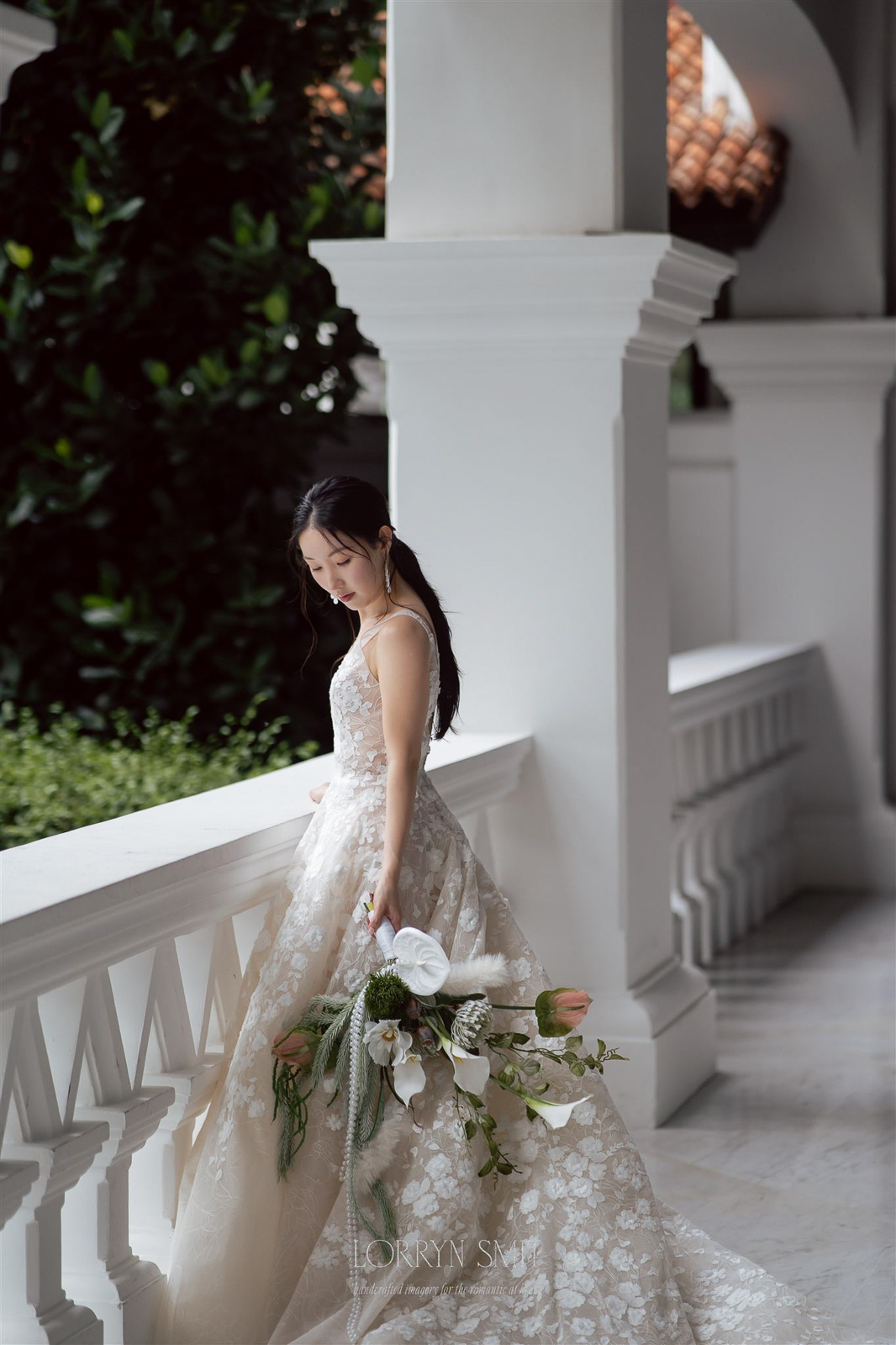 A woman in a white floral wedding dress stands by a white railing, holding a bouquet of flowers, with greenery and architectural columns evoking the elegance found at top Asia wedding destinations.