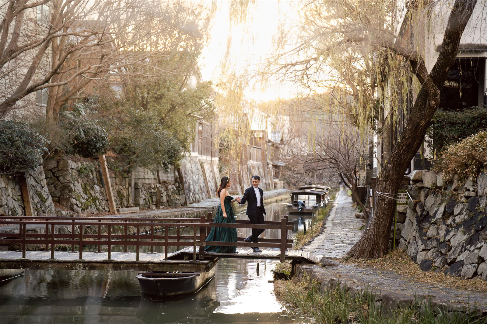 A couple dressed formally walks across a small wooden bridge over a canal at sunset, surrounded by stone walls, trees, and moored boats.