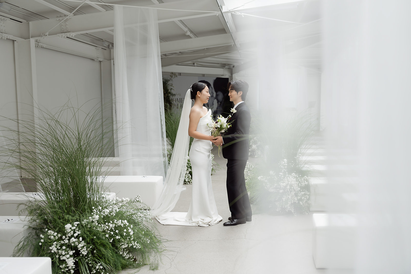 A bride and groom stand facing each other in a bright, modern indoor venue at Aube Studio in Seoul, surrounded by white decor and elegant green floral arrangements, creating a stunning Gallery Wedding atmosphere.