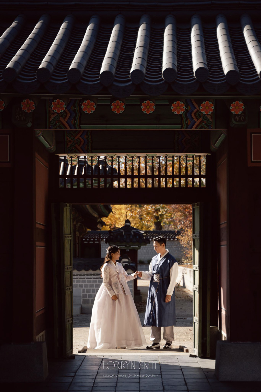 A woman and man in traditional Korean hanbok stand facing each other under a wooden gate at a historic building, with autumn trees in the background—an enchanting scene that highlights Korea among top Asia wedding destinations.