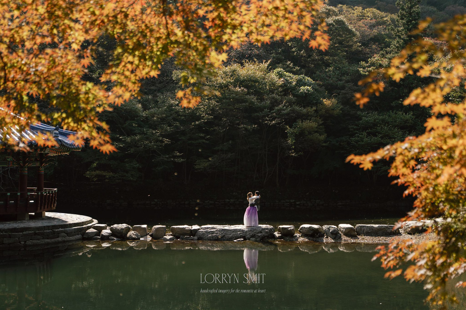 A person in traditional clothing stands on a stone path beside a calm pond, surrounded by trees with autumn foliage—evoking the enchanting beauty of top Asia wedding destinations.