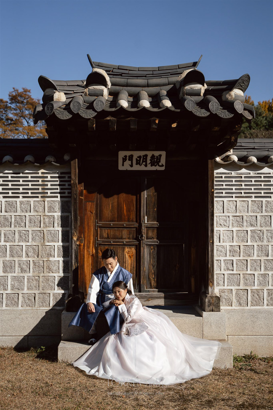 A couple dressed in traditional Korean hanbok sit together in front of a wooden gate with stone walls, under clear sky.