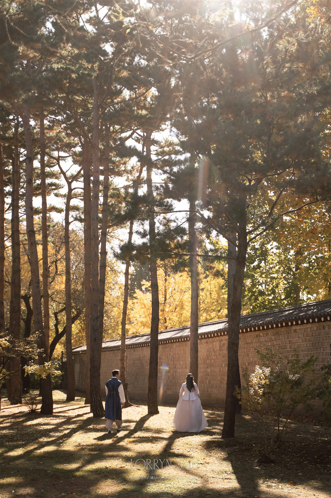 Two people in traditional Korean clothing walk side by side under tall trees near a stone wall on a sunny day, capturing the timeless beauty of one of Asia's top wedding destinations.