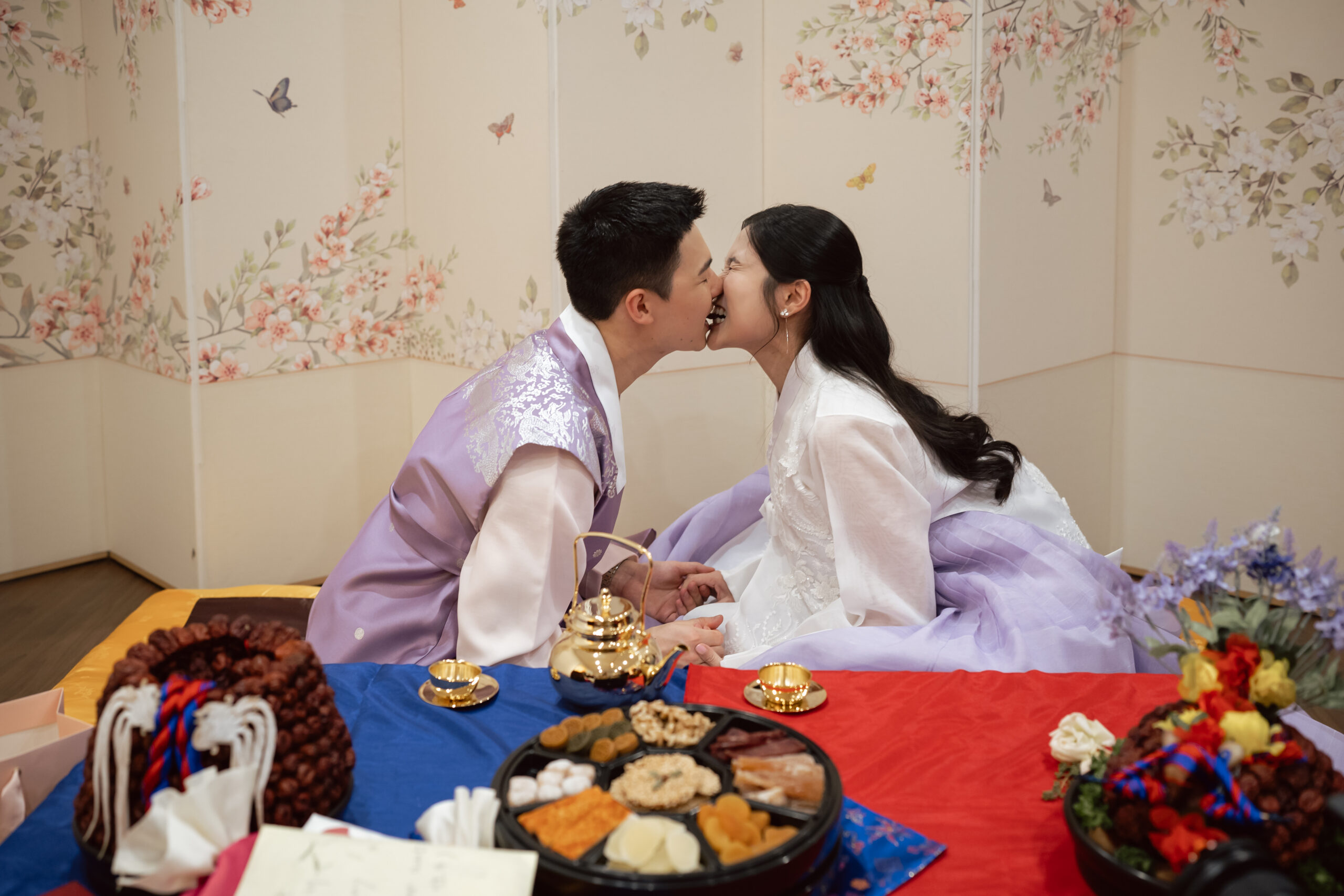 A couple dressed in traditional Korean attire shares a kiss while seated at a table set with ceremonial food, beautifully capturing Korea wedding traditions against a decorative floral screen.