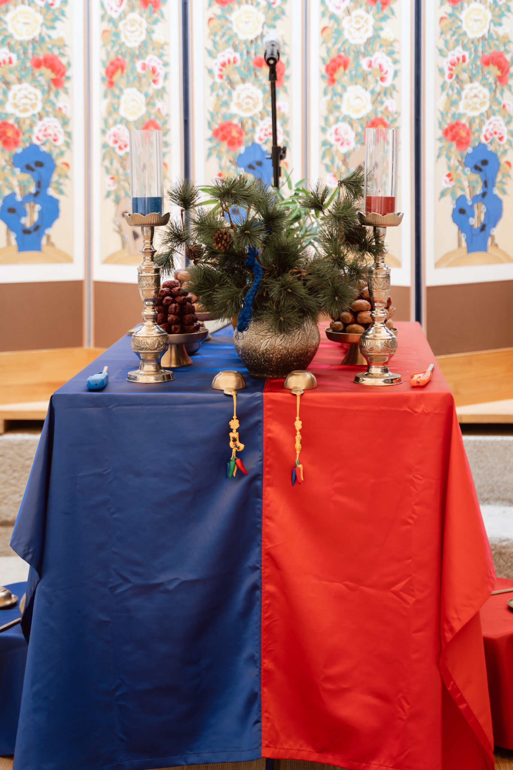 A table covered with blue and red cloths—evoking Korea wedding traditions—displays candles, nuts, a pine branch arrangement, and traditional ornaments; a decorative folding screen stands behind.