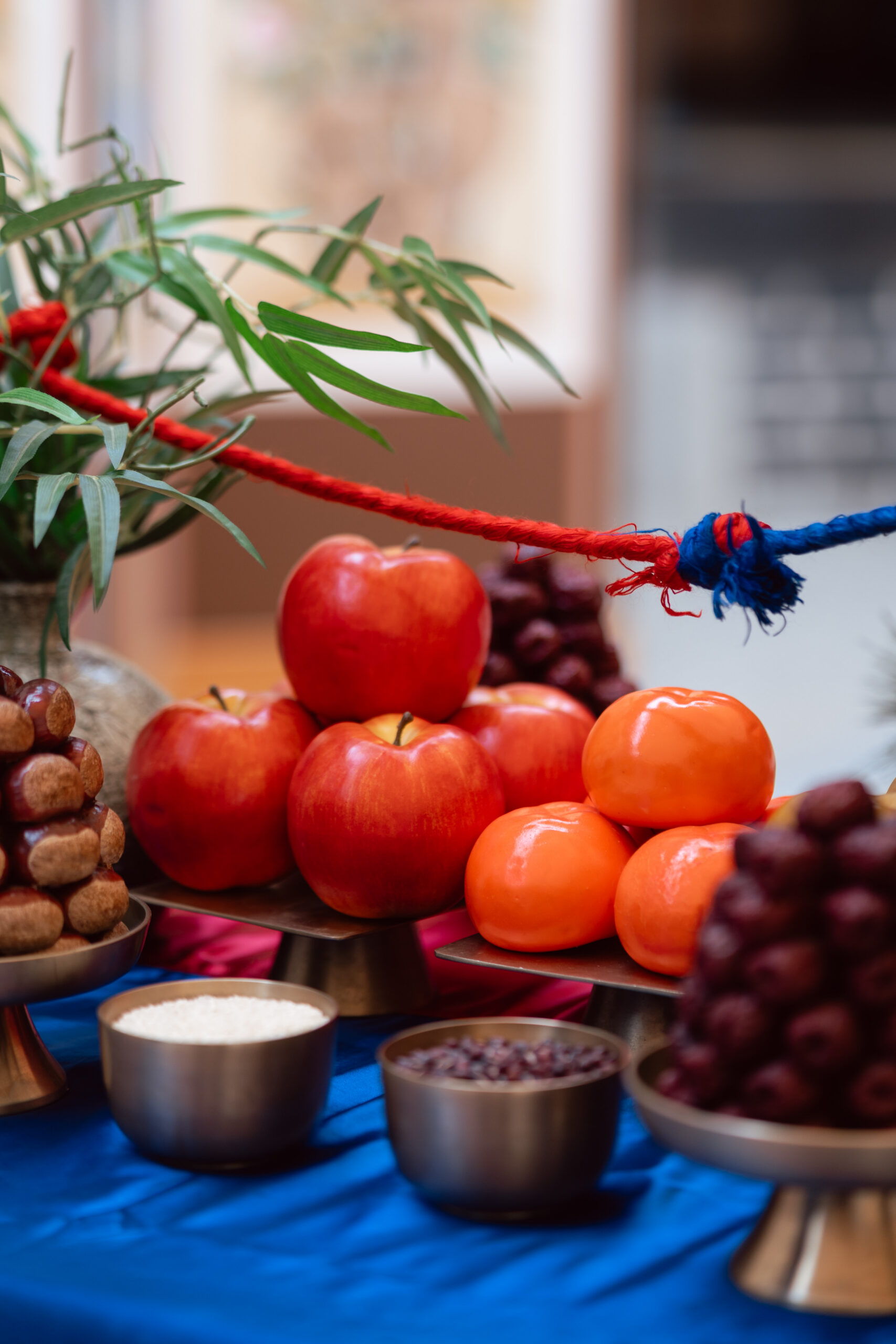 A table displays apples, persimmons, grapes, and small round pastries on trays, with bowls of rice and beans—arranged with greenery and a red and blue rope, reflecting elements of Korea wedding traditions.