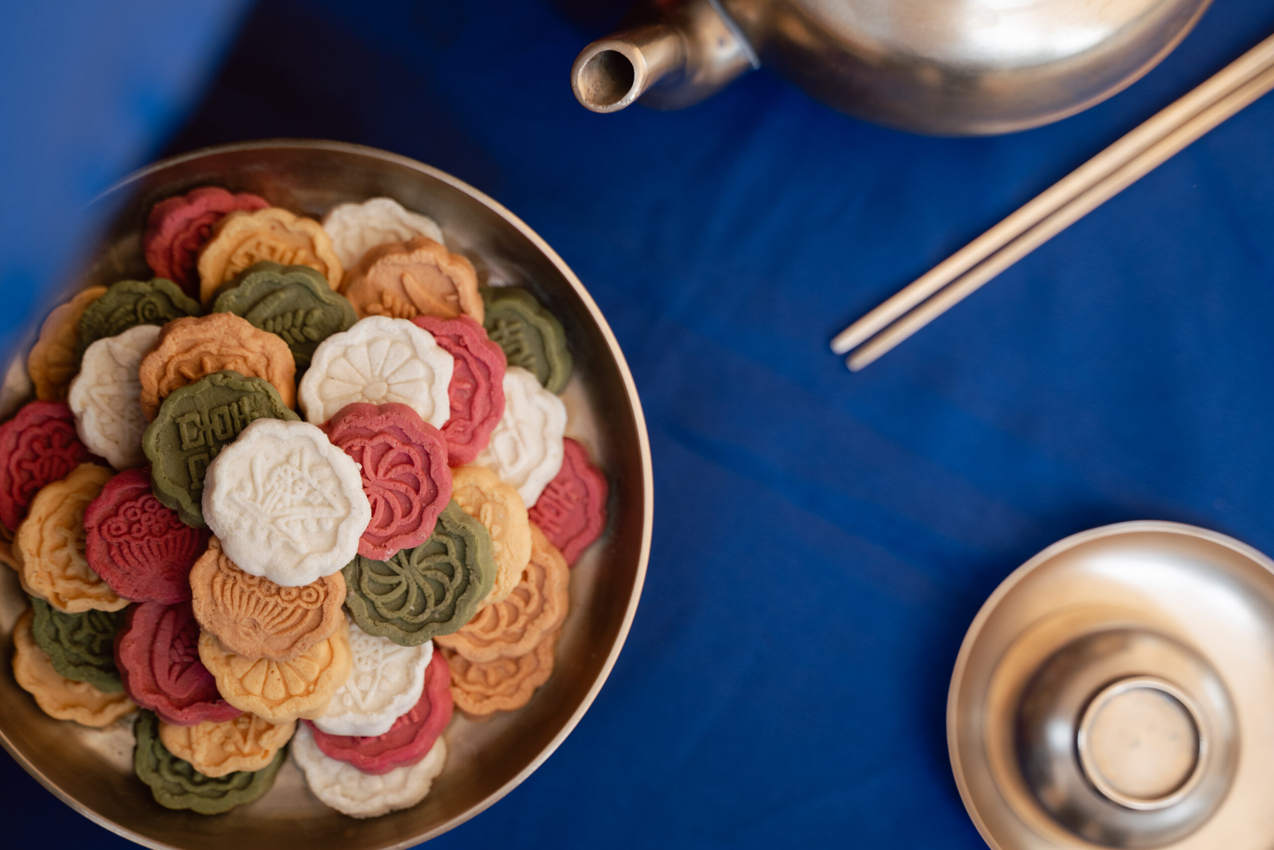 A bowl of colorful flower-shaped cookies sits next to a teapot, chopsticks, and a metal cup on a blue tablecloth, evoking the elegance of Korea wedding traditions.