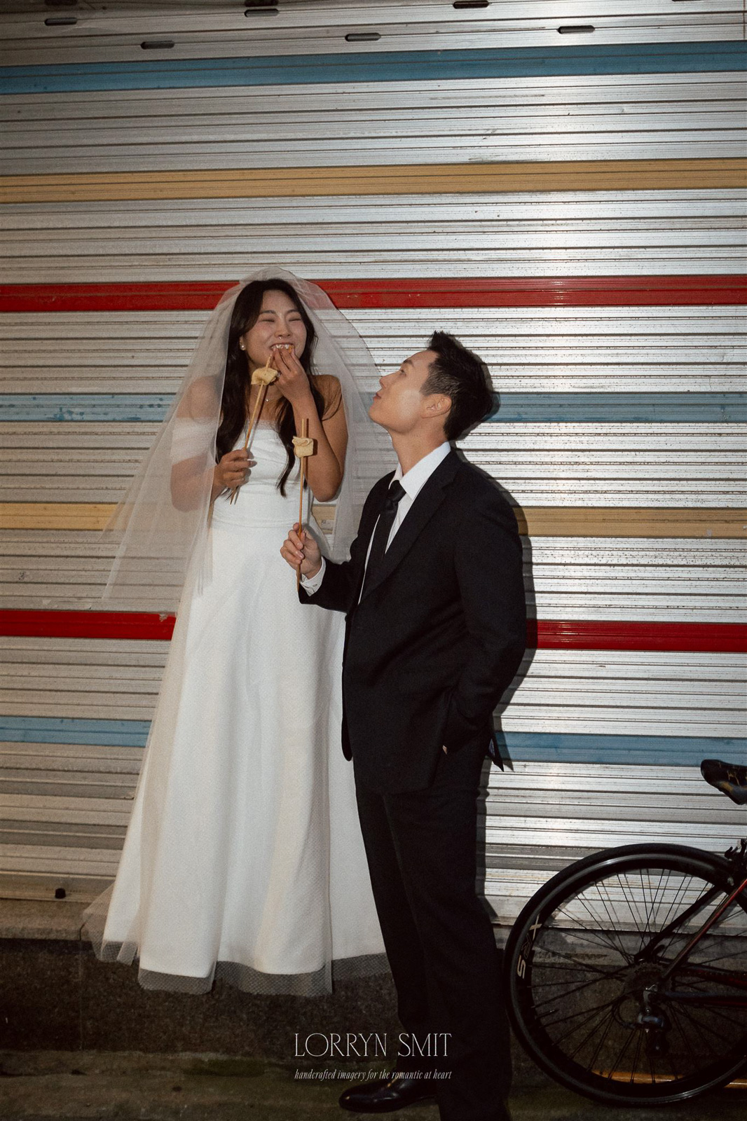 During an autumn pre wedding shoot, a bride in a white dress and veil enjoys a snack with a man in a suit beside her, both holding food, standing in front of a striped metal wall next to a bicycle.