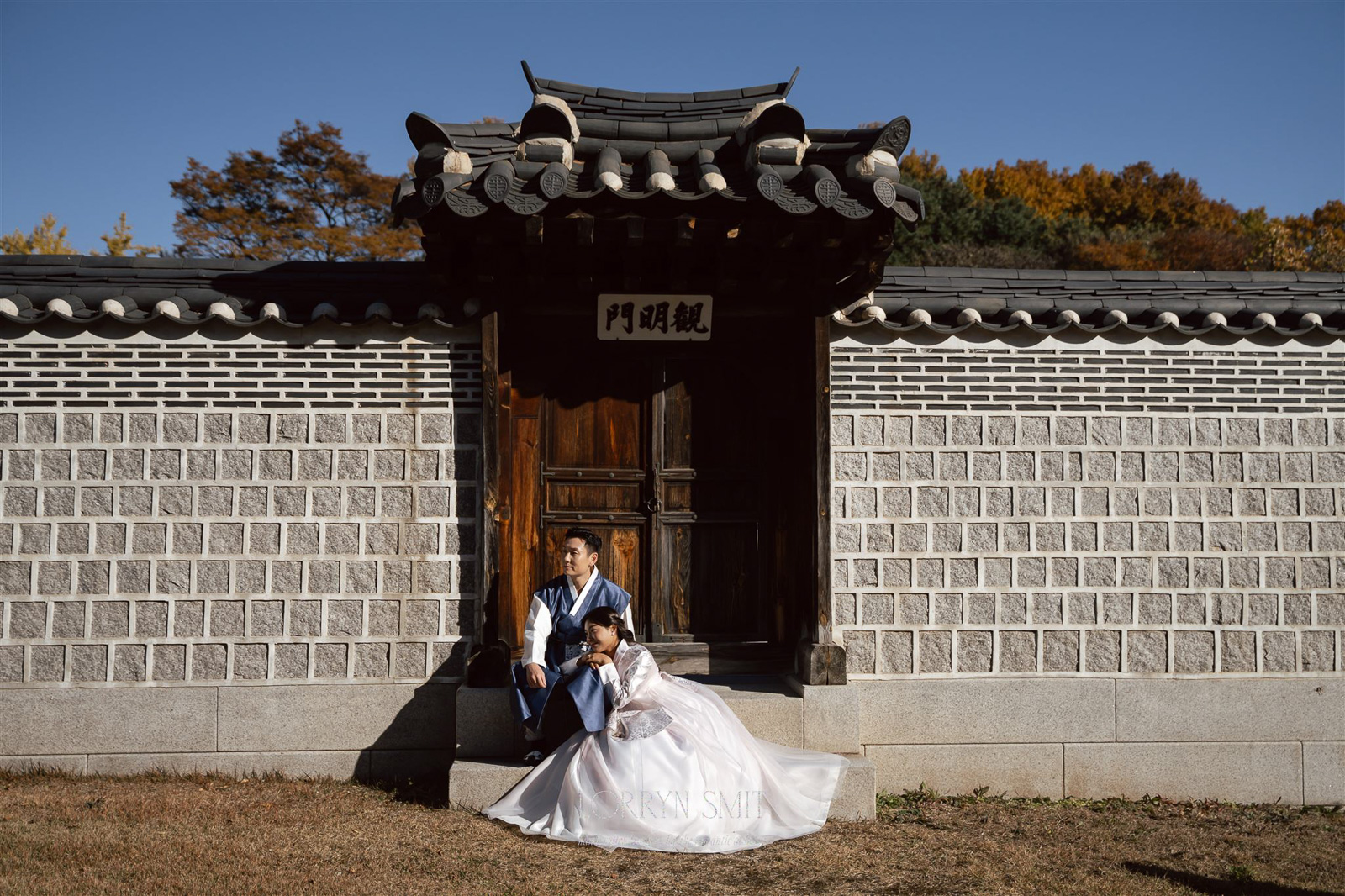 A couple in traditional Korean attire sits in front of a wooden gate set in a stone wall, capturing an autumn pre wedding shoot with colorful trees glowing in the background.