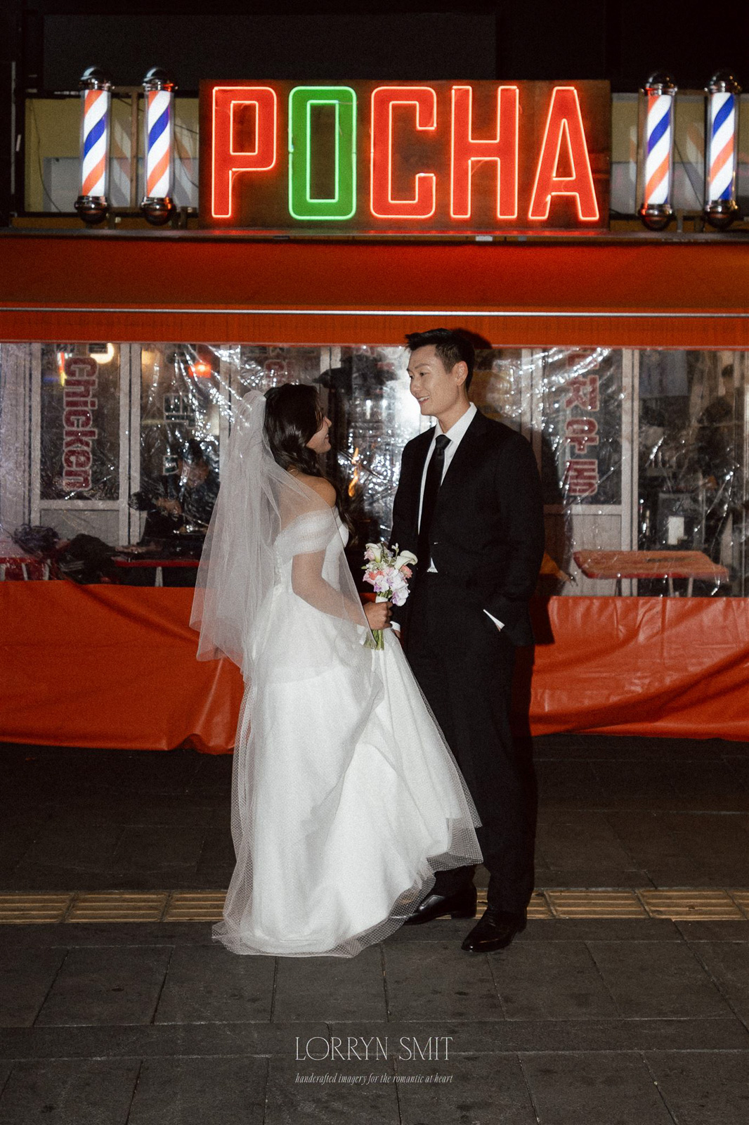 A bride and groom stand facing each other at night, dressed in wedding attire for an autumn pre wedding shoot, in front of a neon-lit sign reading "POCHA".