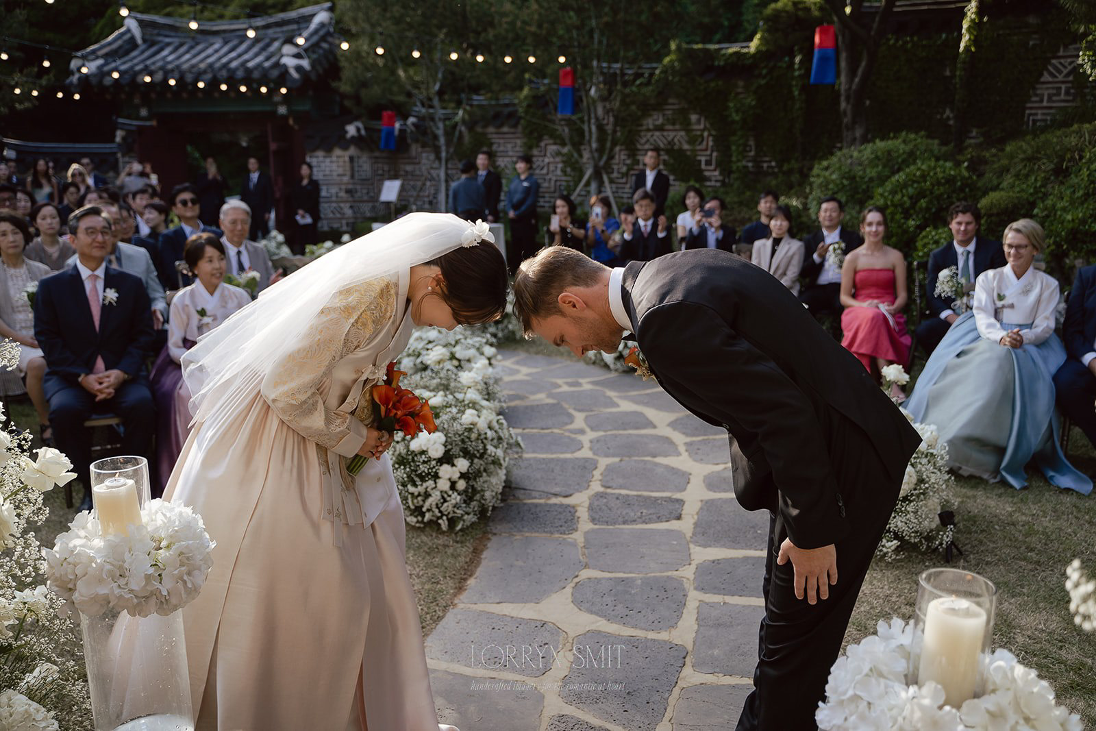 A bride and groom bow to each other during an outdoor wedding ceremony, surrounded by guests and white floral decorations.