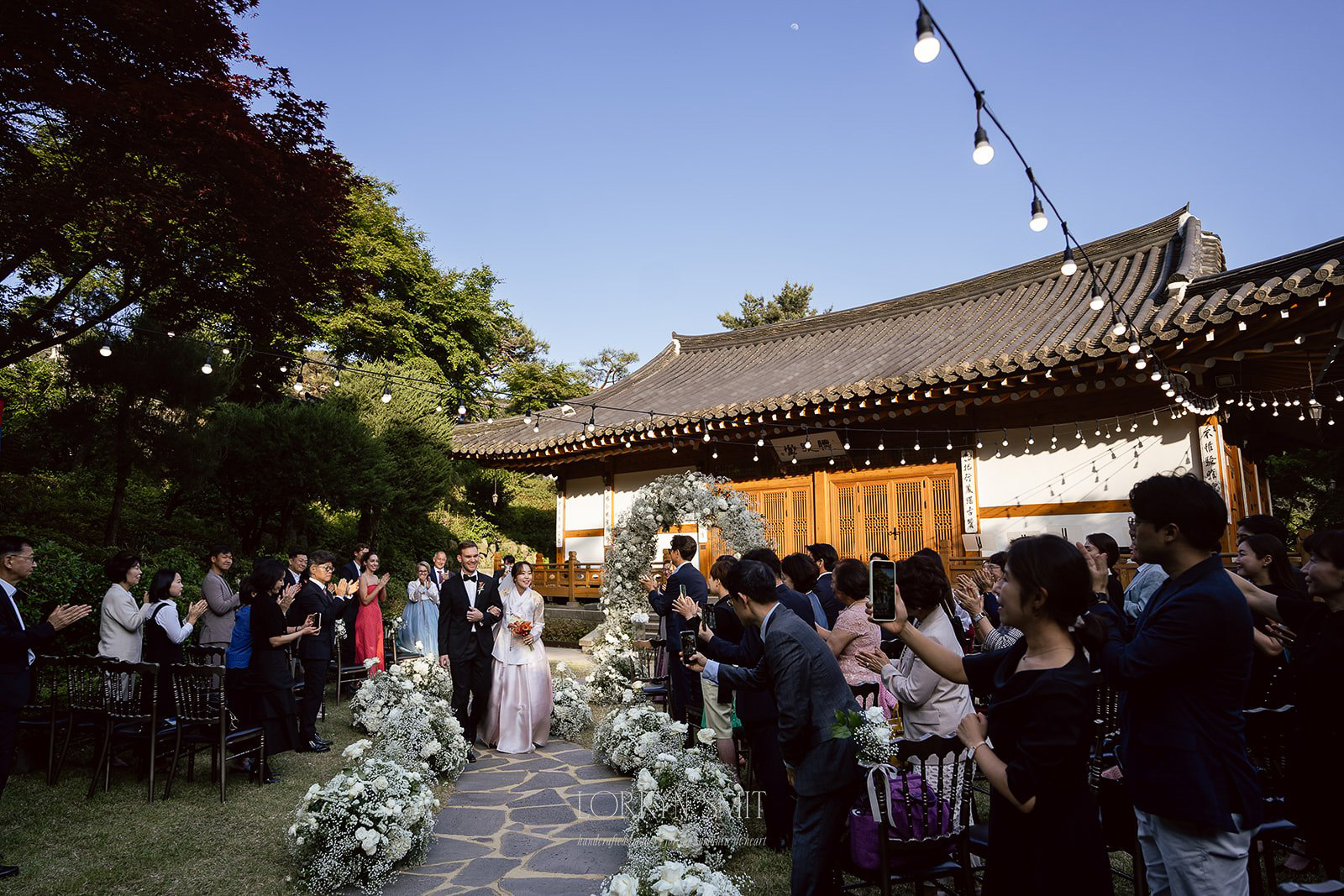 A bride walks down an outdoor aisle lined with flowers, surrounded by guests, at a traditional-style building under string lights.
