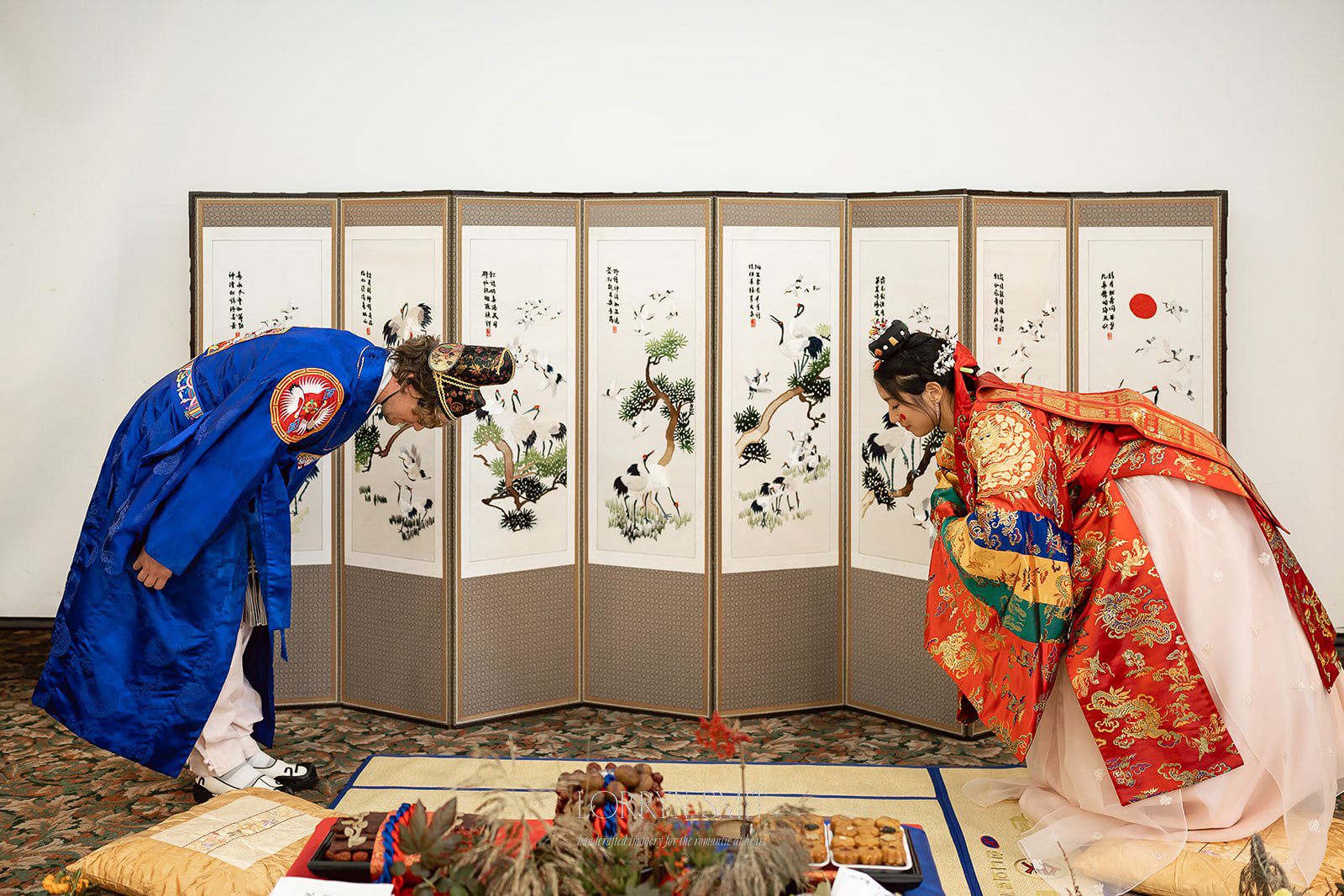 Two people in traditional Korean wedding attire bow to each other in front of a decorative folding screen during a ceremony, one of many wedding traditions. 