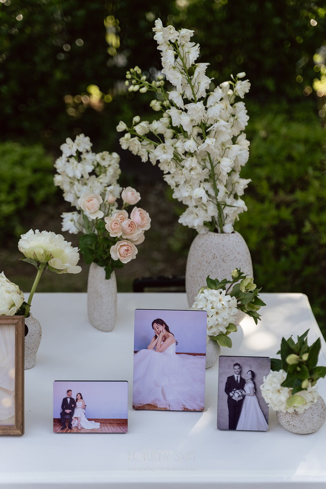 A white table displays framed wedding photos and is decorated with vases of white and light pink flowers against a green outdoor background, part of the wedding traditions in Korea.
