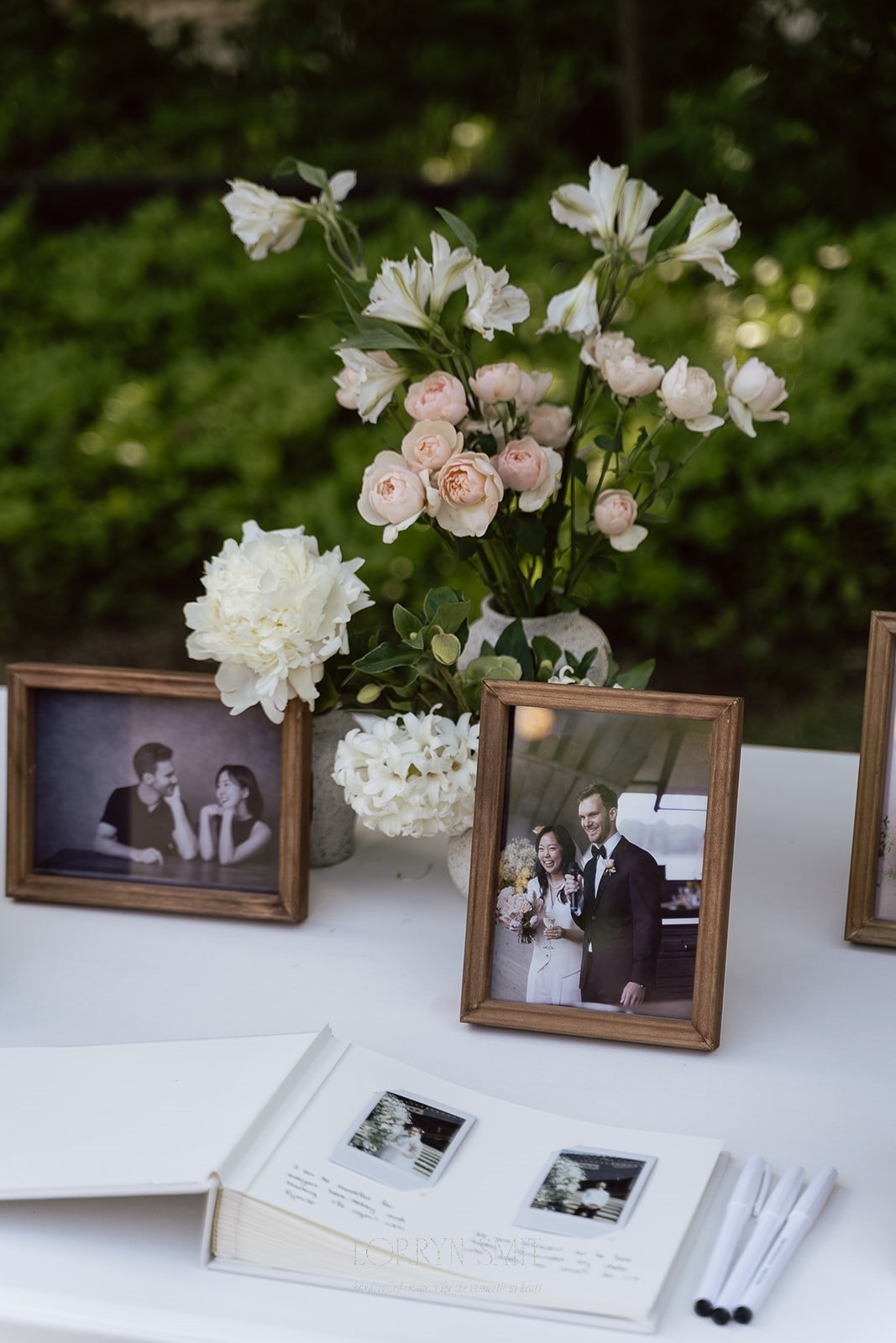A table with framed photos, a bouquet of flowers, a guestbook with instant photos, and pens arranged neatly on top.