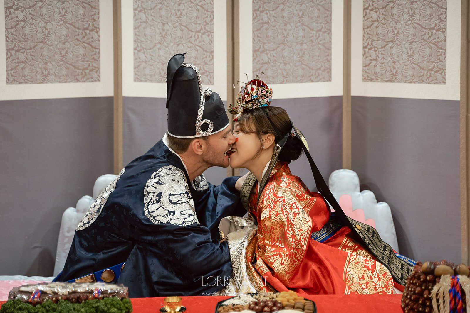 A couple in Korea during a Paebaek ceremony wearing Hanbok wedding attire lean in for a kiss while seated at a decorated table during a ceremony.