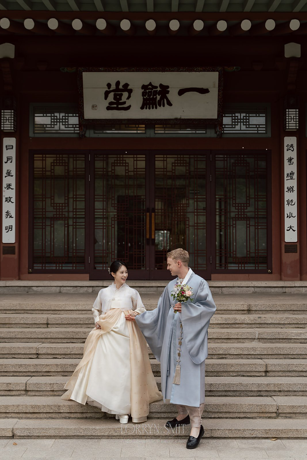 A couple in traditional Korean hanbok stands on temple steps, holding hands and looking at each other. The man holds a bouquet; Korean writing is visible on the building behind them.