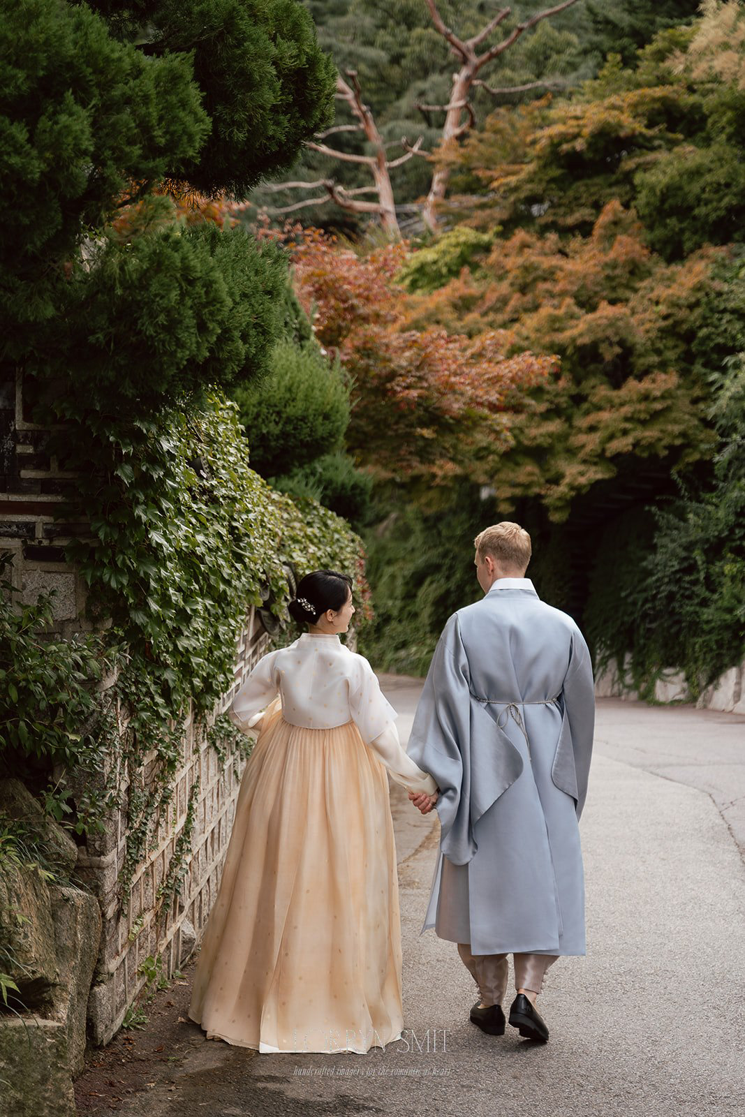 A couple dressed in traditional Korean hanbok walk hand in hand along a tree-lined path.
