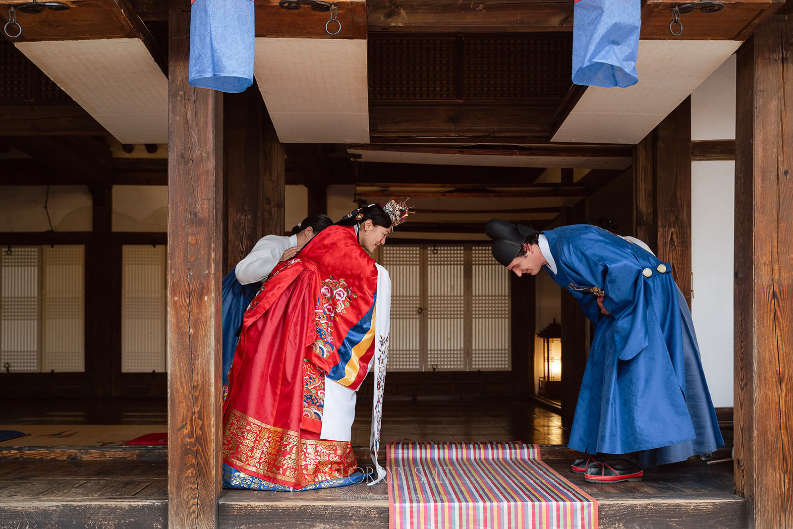 Two people in traditional Korean hanbok bow to each other inside a wooden building, participating in a formal ceremony.