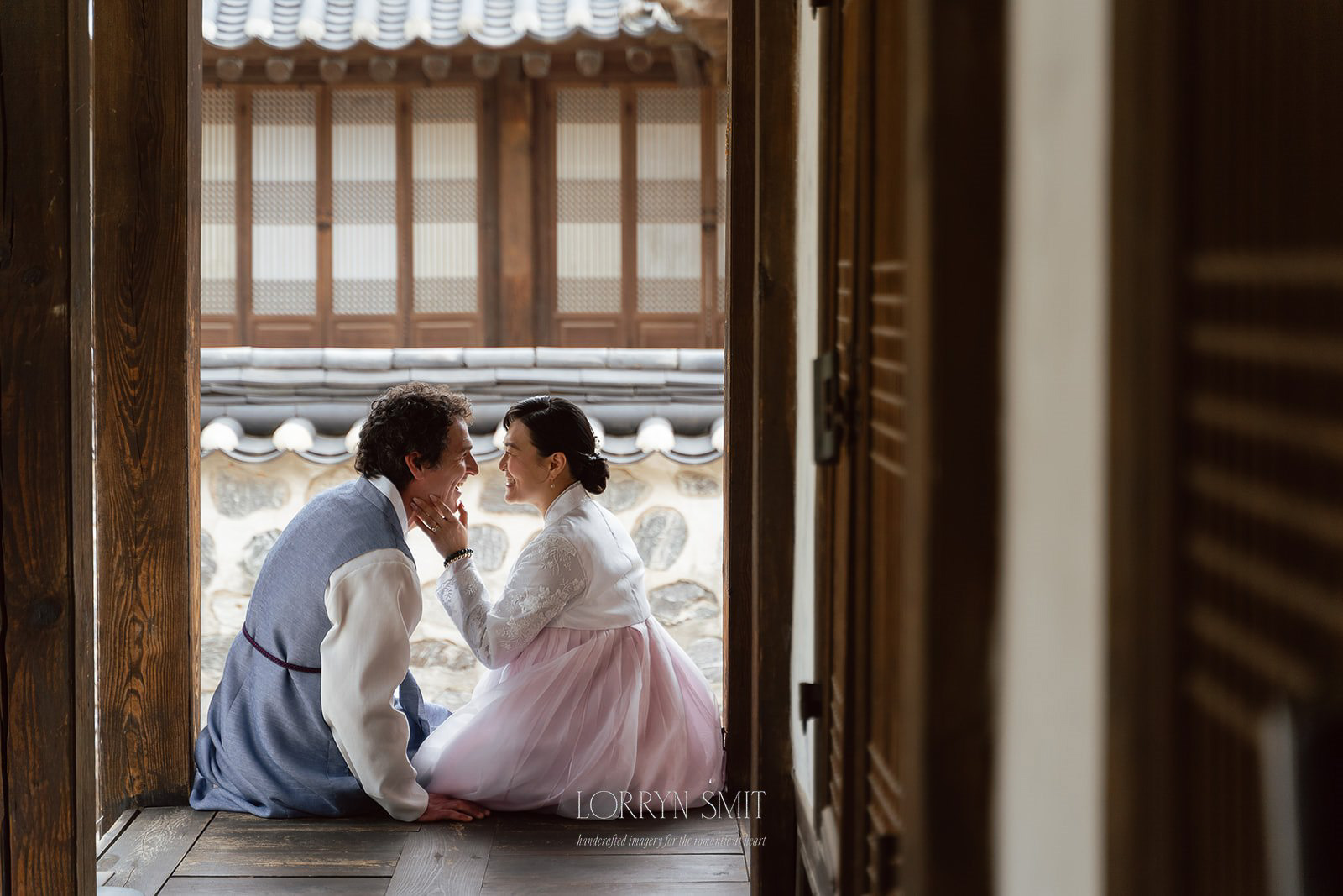 A couple in Korea wearing Hanbok, one of many Korean wedding traditions, sit closely together on a wooden porch, smiling at each other in front of a traditional building.