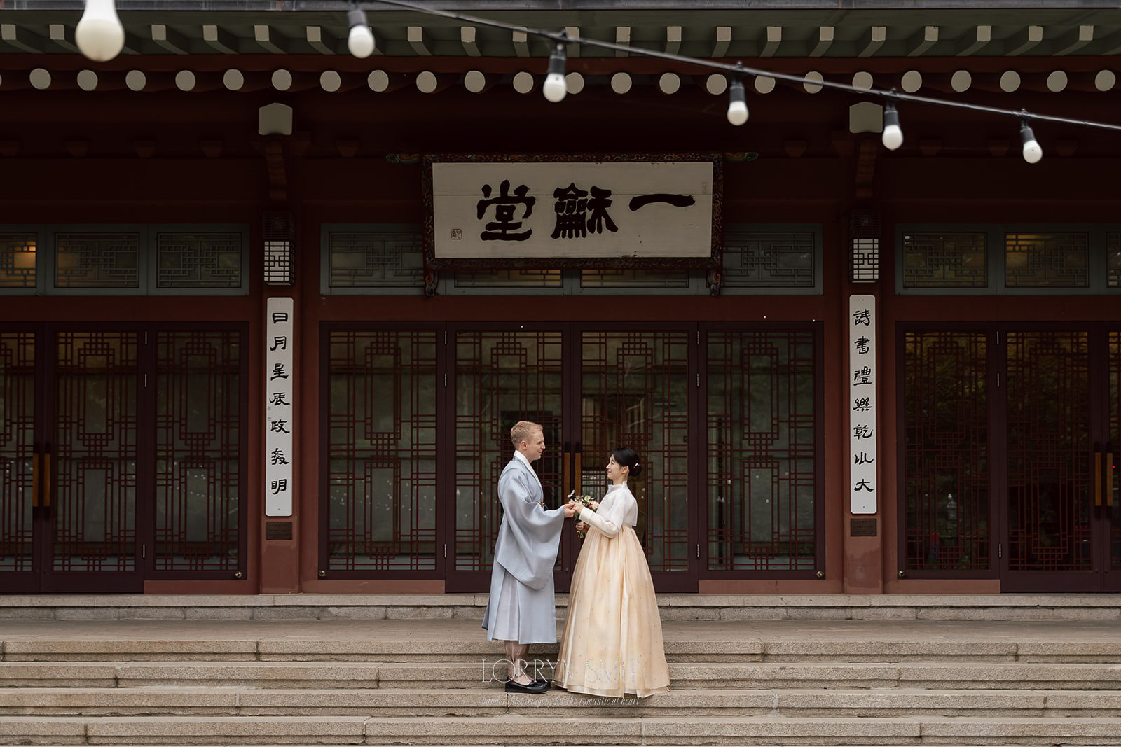 A couple in traditional Korean hanbok stands facing each other on steps in front of a historical building with Korean signage.