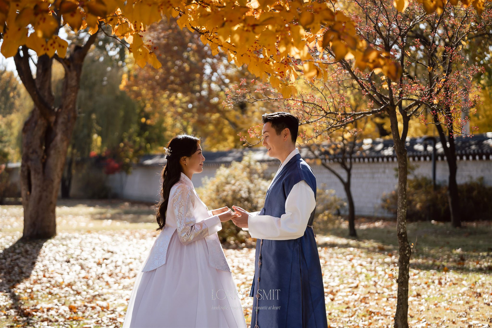 A couple in traditional Korean attire stands facing each other, holding hands in a sunlit park with autumn foliage, capturing a romantic autumn pre wedding shoot.