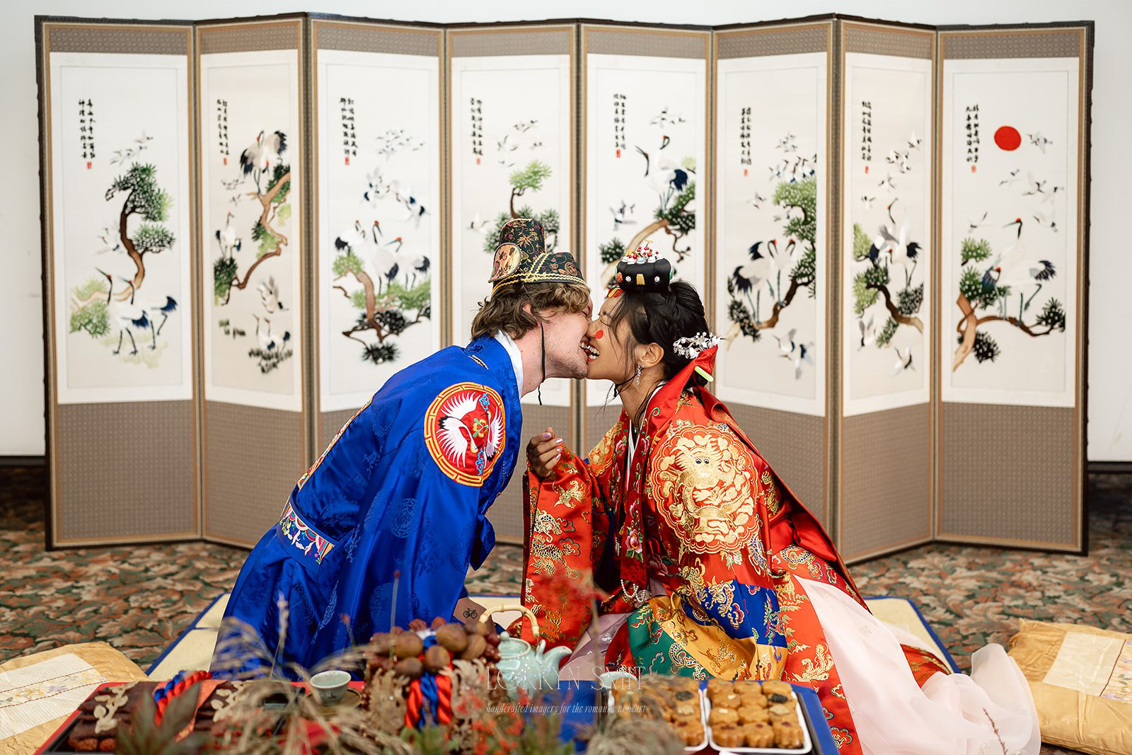 A couple in traditional Korean wedding attire performs a ritual in front of a decorative folding screen, kneeling and touching their noses together.
