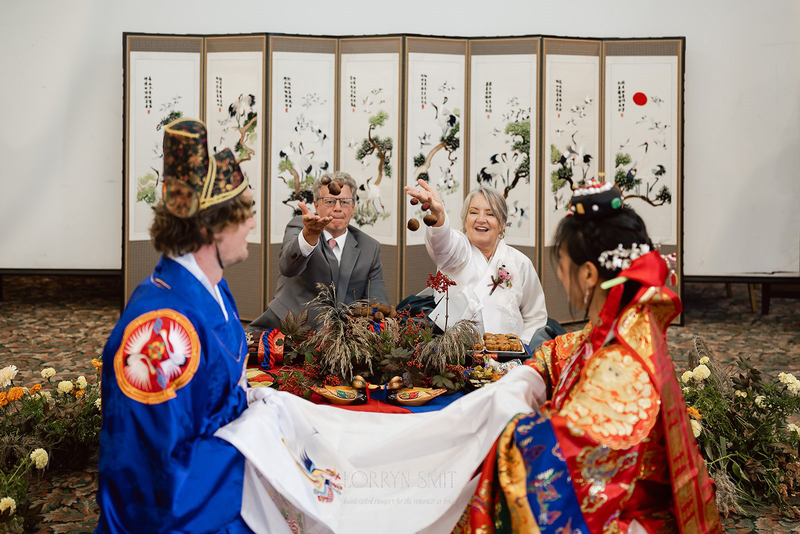 Two couples in traditional and modern attire participate in a Korean wedding ceremony, seated in front of a decorated table with food and a floral folding screen.