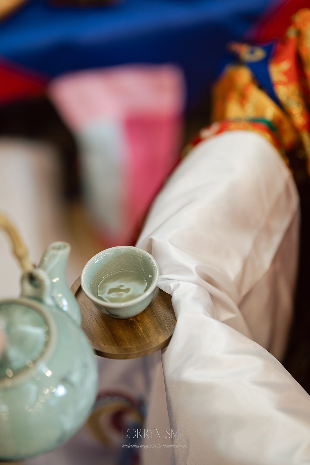 A person in traditional attire holds a wooden tray with a ceramic teacup and teapot, preparing to serve tea.