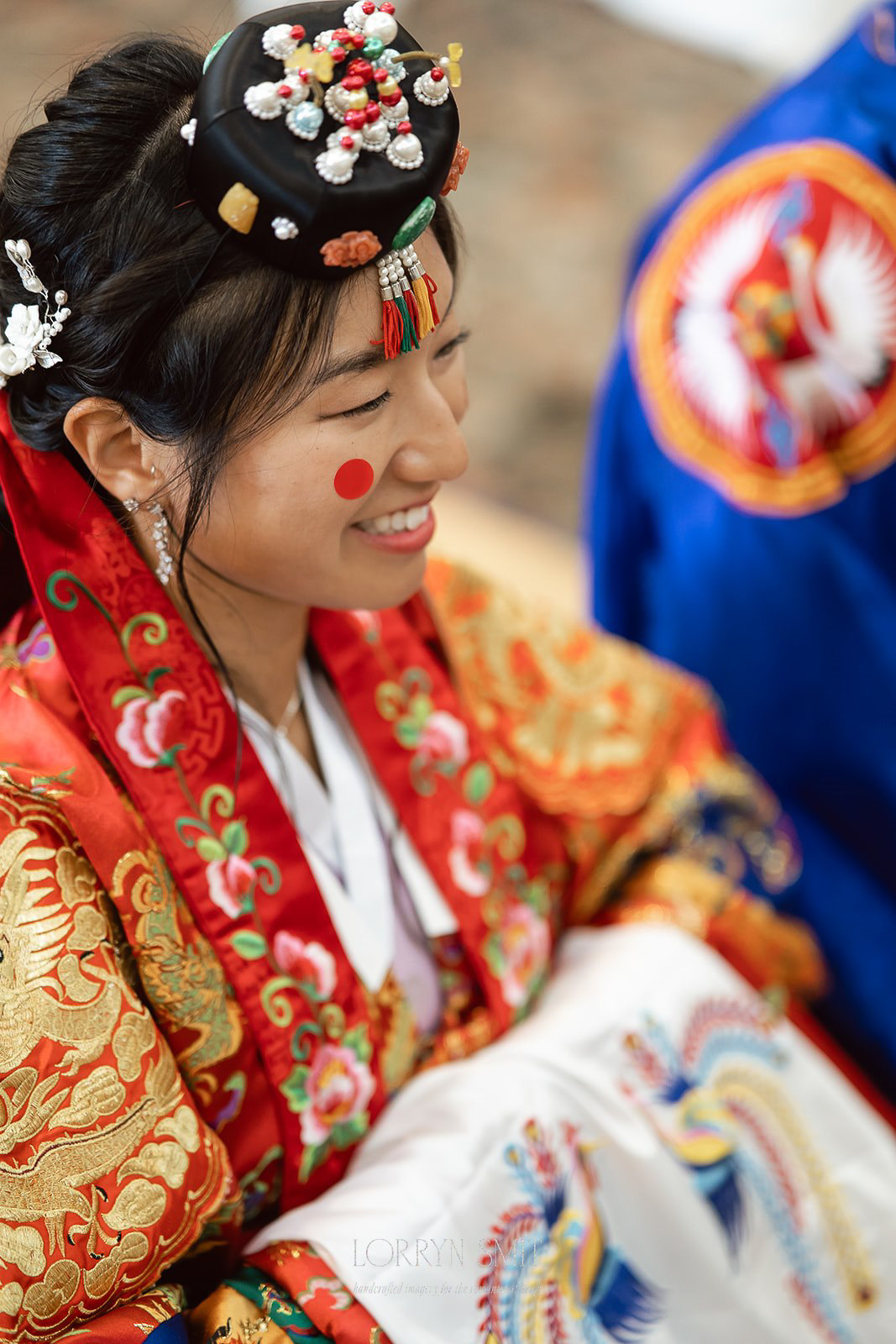 A woman wearing traditional Korean wedding attire with a red and gold hanbok and headpiece, smiling and seated.