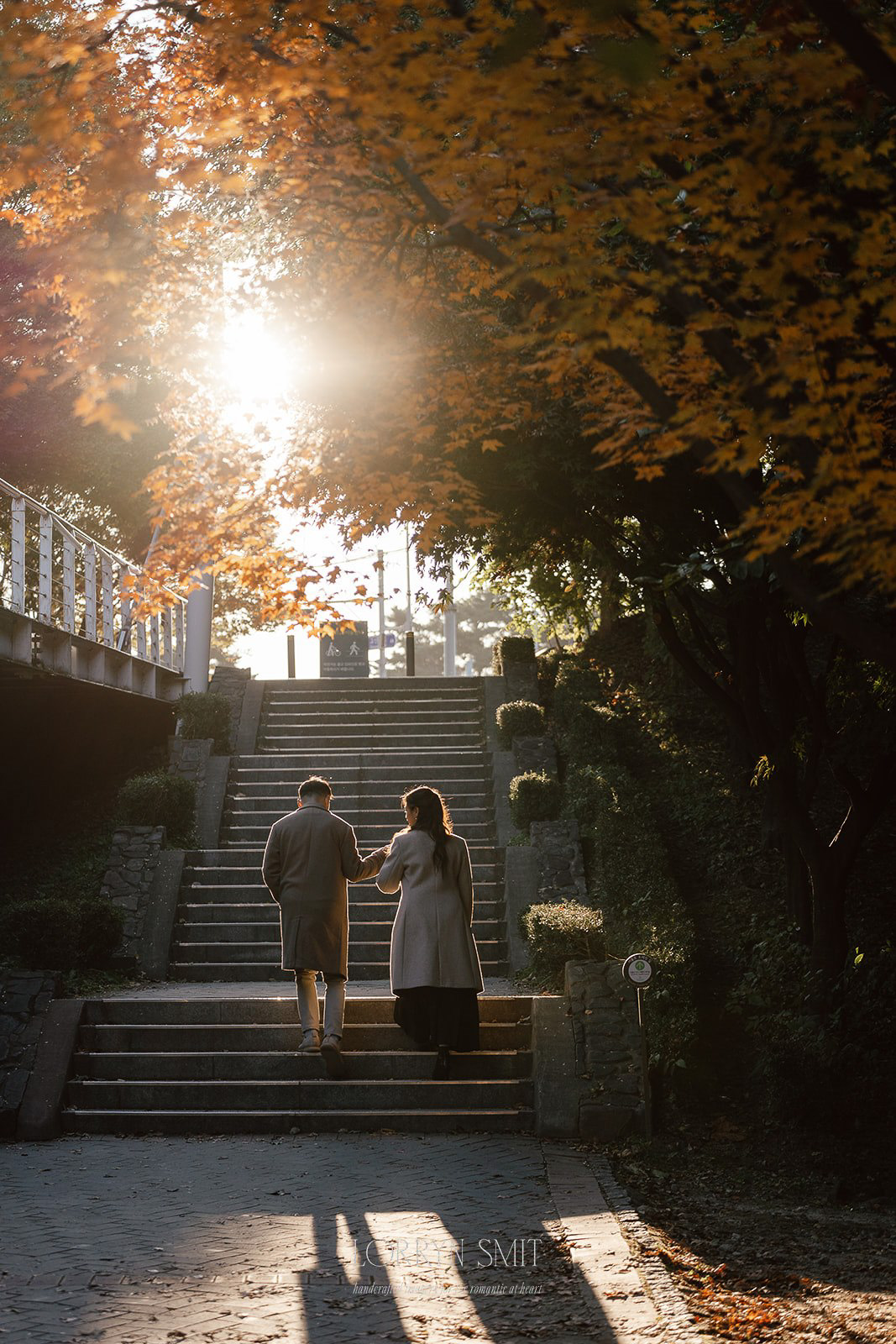 A couple walks up stone steps in a park, holding hands, with sunlight filtering through autumn trees during their Korea pre wedding.