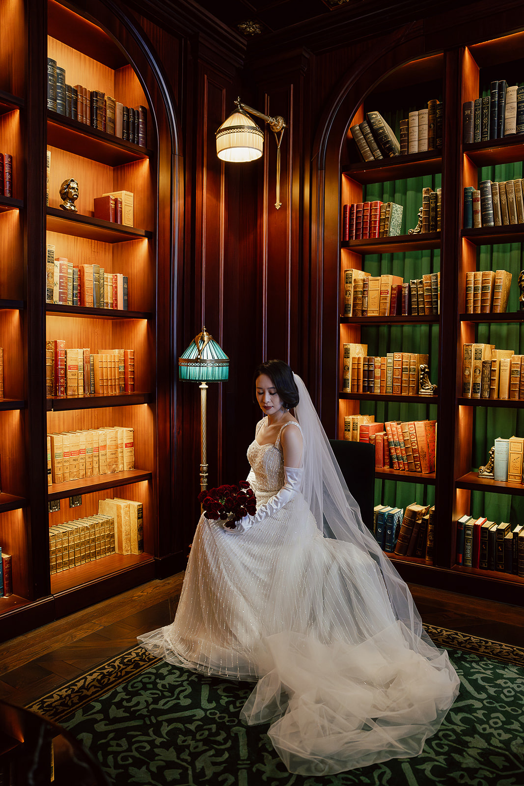 A bride in a white wedding dress and veil sits holding a bouquet in a wood-paneled library with shelves filled with books.