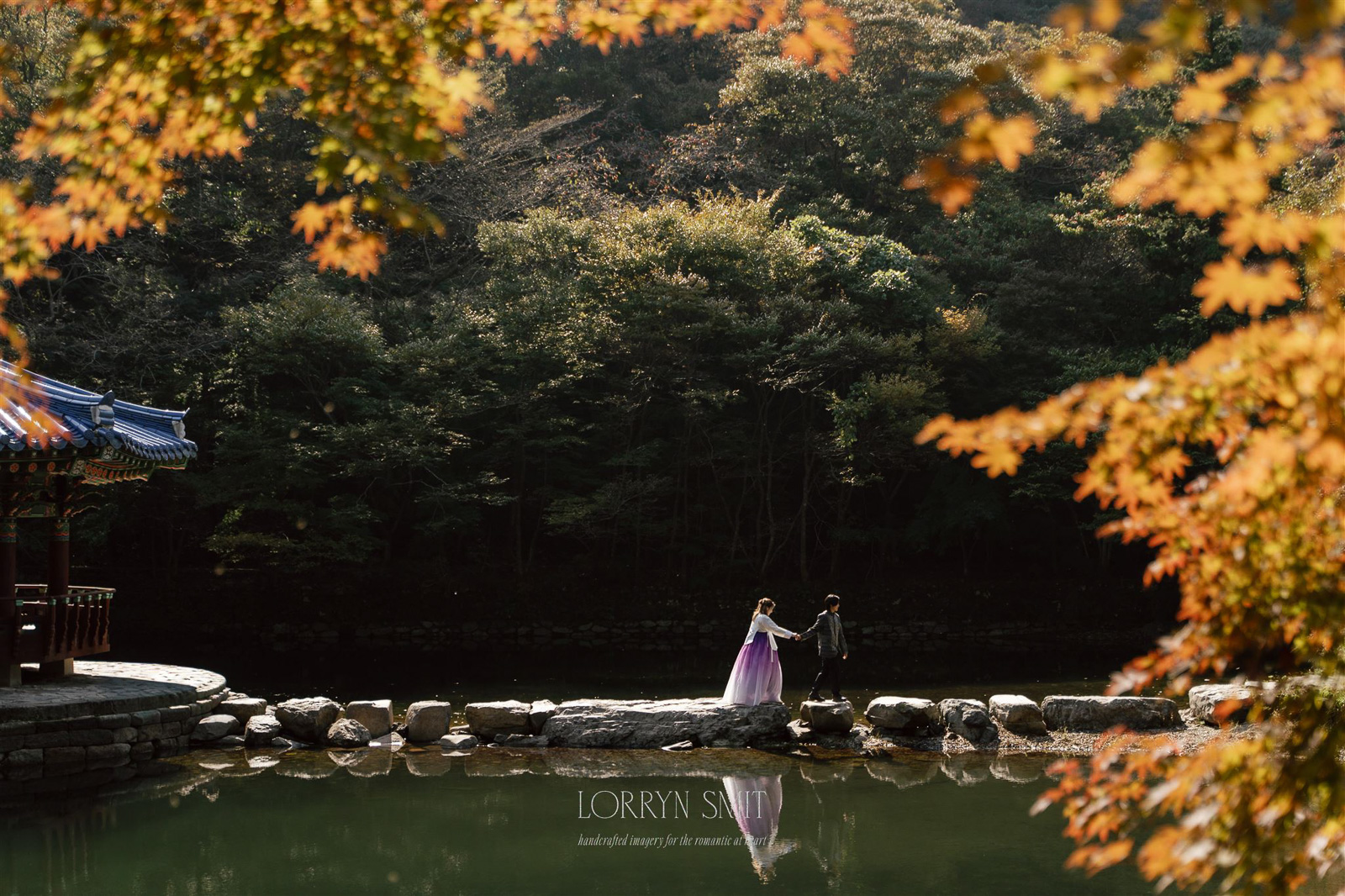 A couple in traditional attire stands on stone steps over a tranquil pond, surrounded by autumn trees, with a pavilion on the left during their Korea pre wedding.