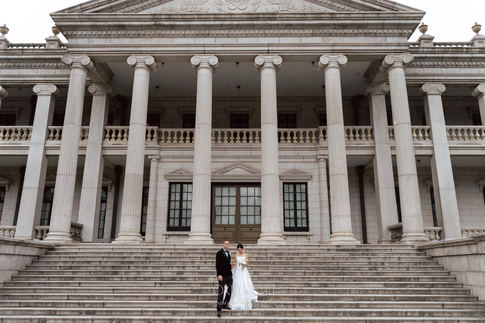 A bride and groom stand together on the steps of a large, neoclassical building with tall columns and ornate architecture during their Korea pre wedding.