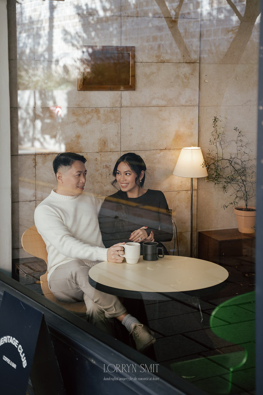 A man and woman sit together at a round table in a cafe, holding hands and smiling, with coffee mugs in front of them during their Korea pre wedding.